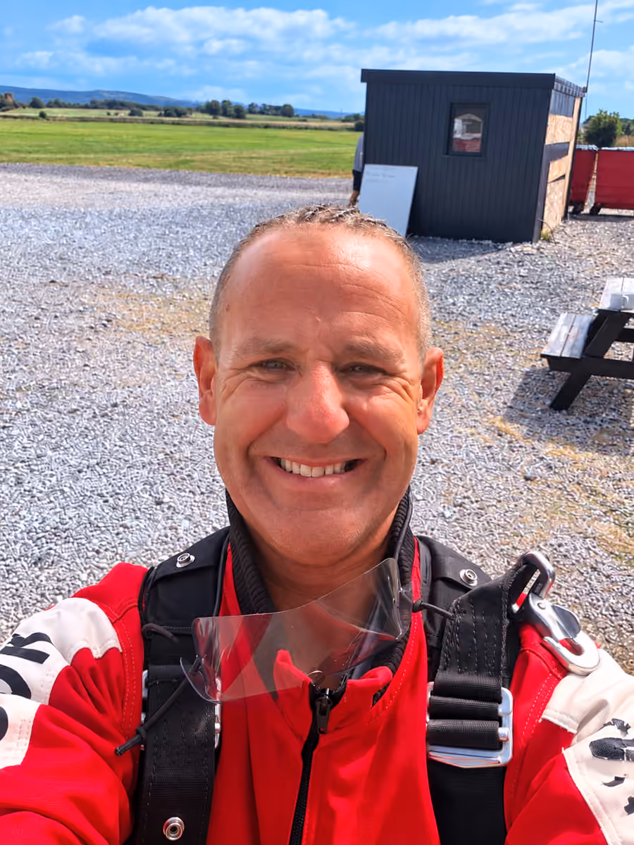 Colin smiling at the airfield in a red skydiving suit and harness.